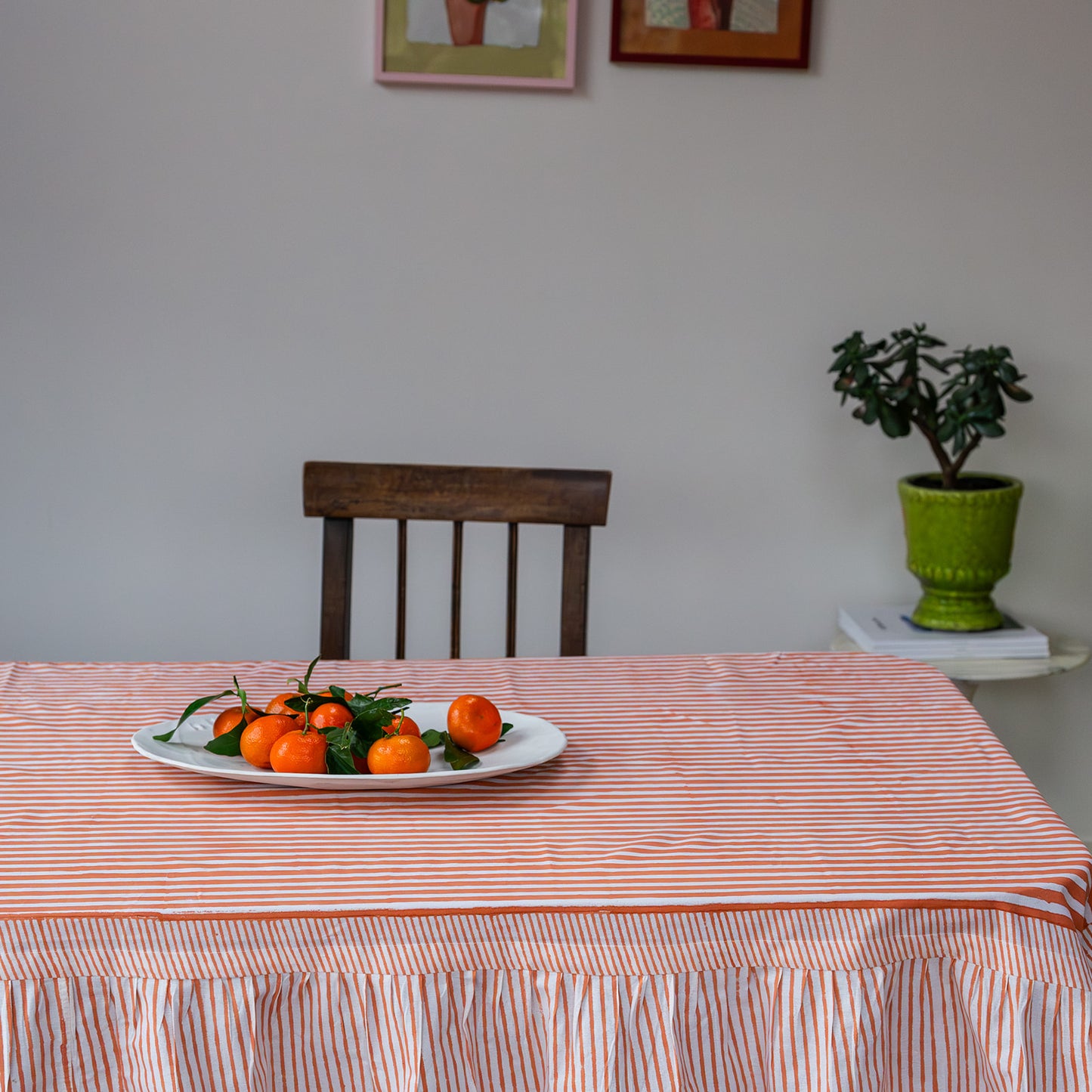 Dining table with a striped tablecloth, plate of oranges, and chair in a room with a plant and framed pictures on the wall.