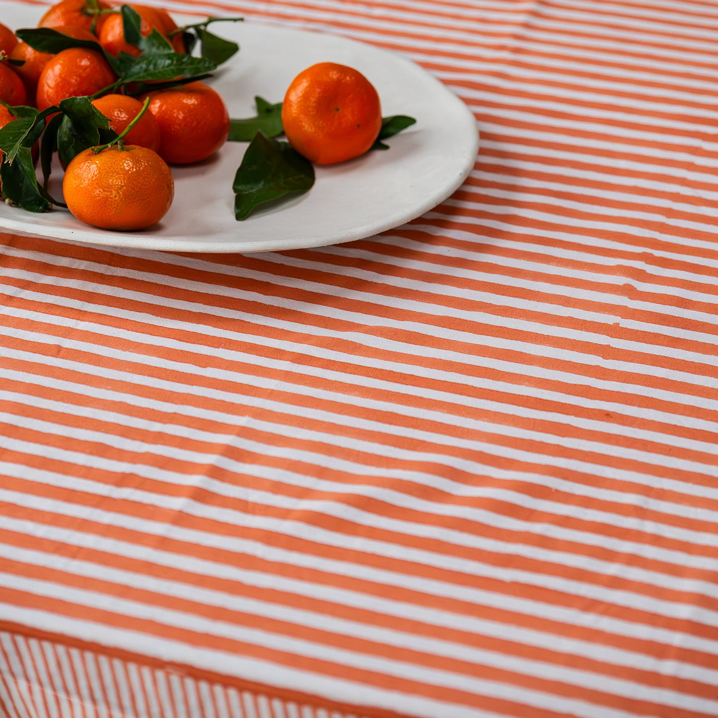 Clementines on a white plate with green leaves on an orange and white striped tablecloth.