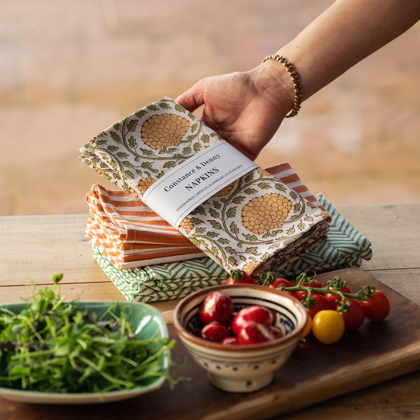 Hand holding a stack of patterned napkins with a blurred background of a table with food.