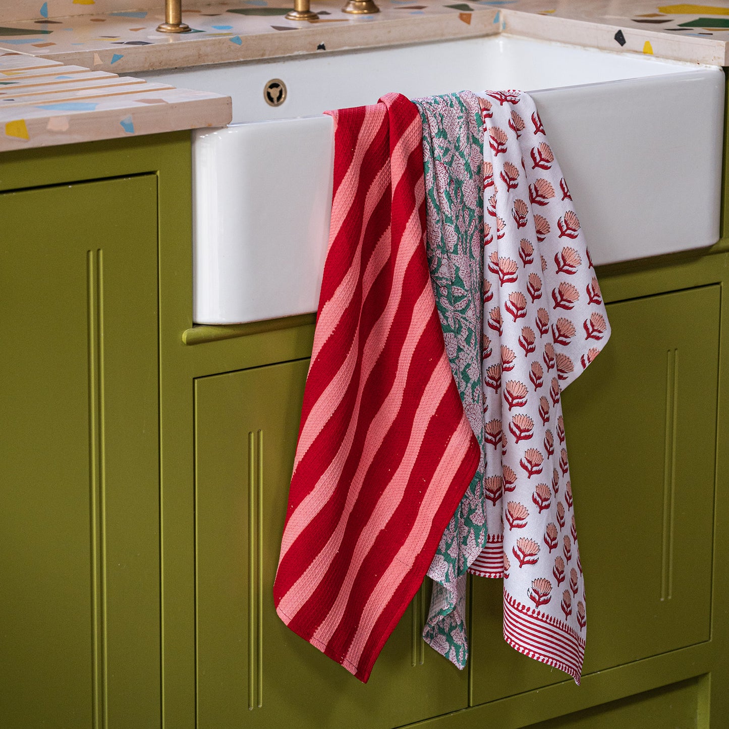 Red striped and patterned towels hanging over a white kitchen sink with green cabinets.