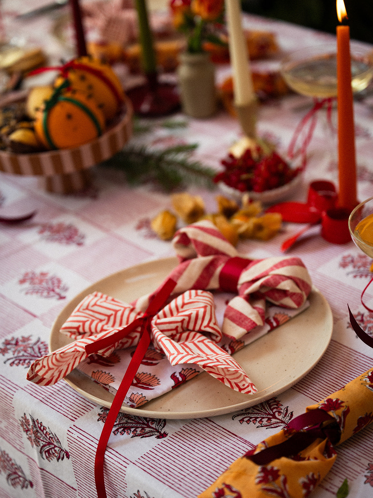 A styled holiday table setting featuring a beige plate with folded napkins in red and white striped and solid red patterns, tied with a red ribbon, on a red-and-white checkered tablecloth.