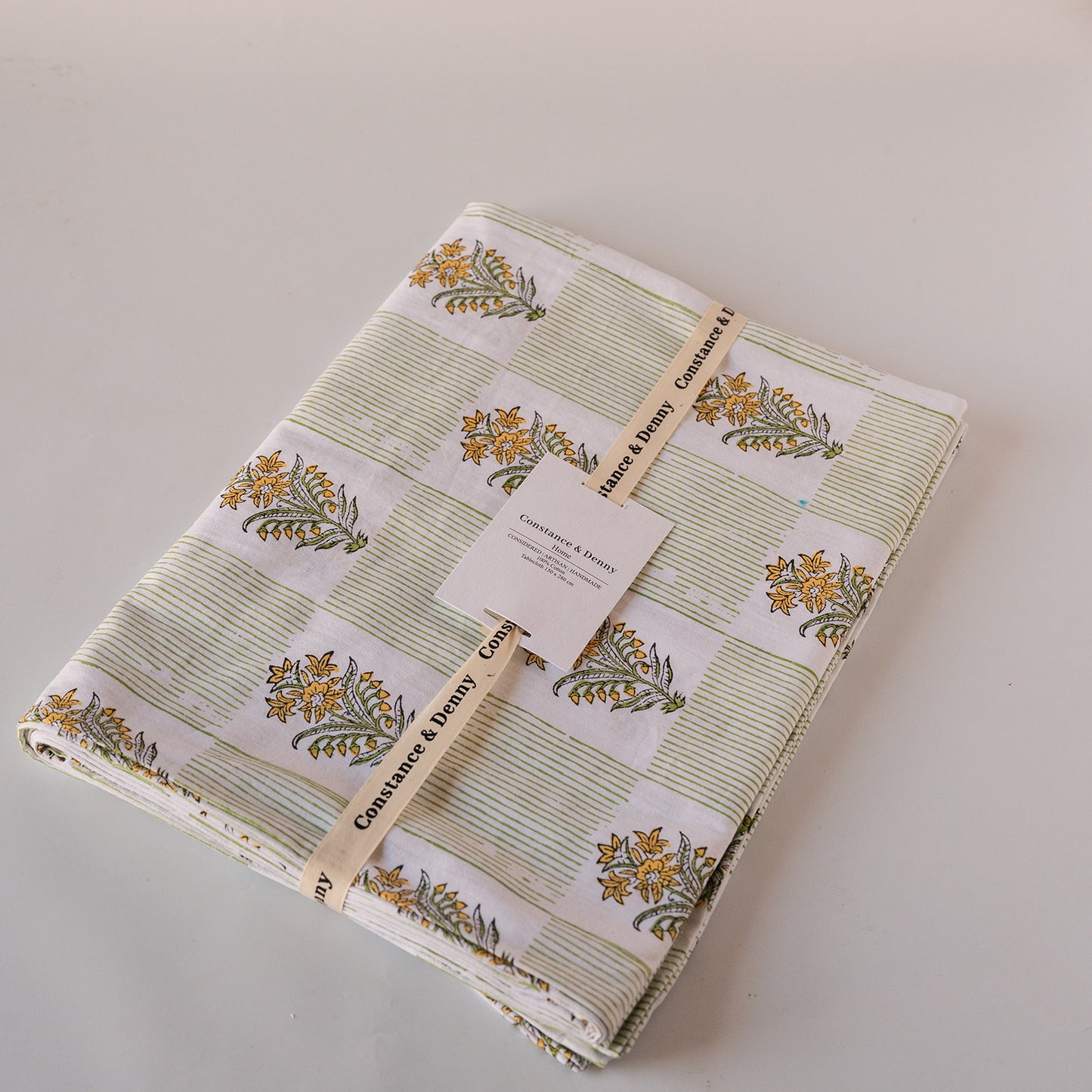 A folded fabric item, likely a quilt or blanket, featuring a patchwork design of green striped squares and white squares with yellow floral motifs. The fabric is wrapped with a paper band and tag, showing the brand 'Coaster & Dolly'.