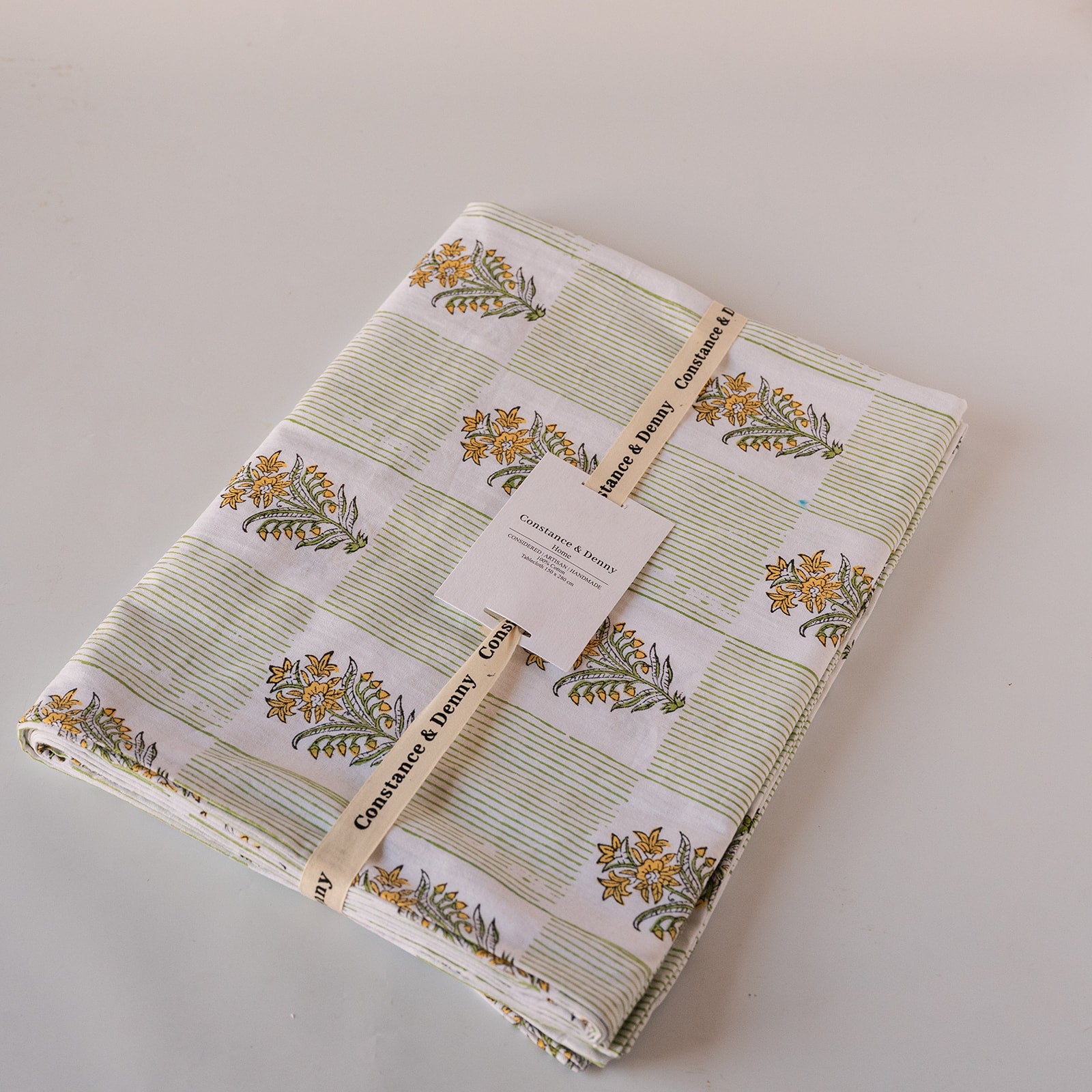 A folded fabric item, likely a quilt or blanket, featuring a patchwork design of green striped squares and white squares with yellow floral motifs. The fabric is wrapped with a paper band and tag, showing the brand 'Coaster & Dolly'.