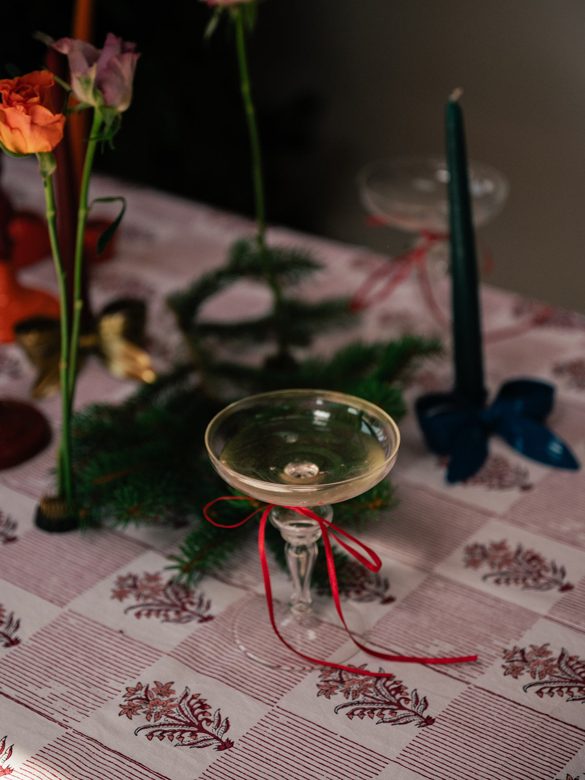 A clear glass coupe-style cocktail glass with a stem, decorated with a red ribbon bow, set on a patterned tablecloth with flowers, greenery, and a dark teal taper candle in the background.