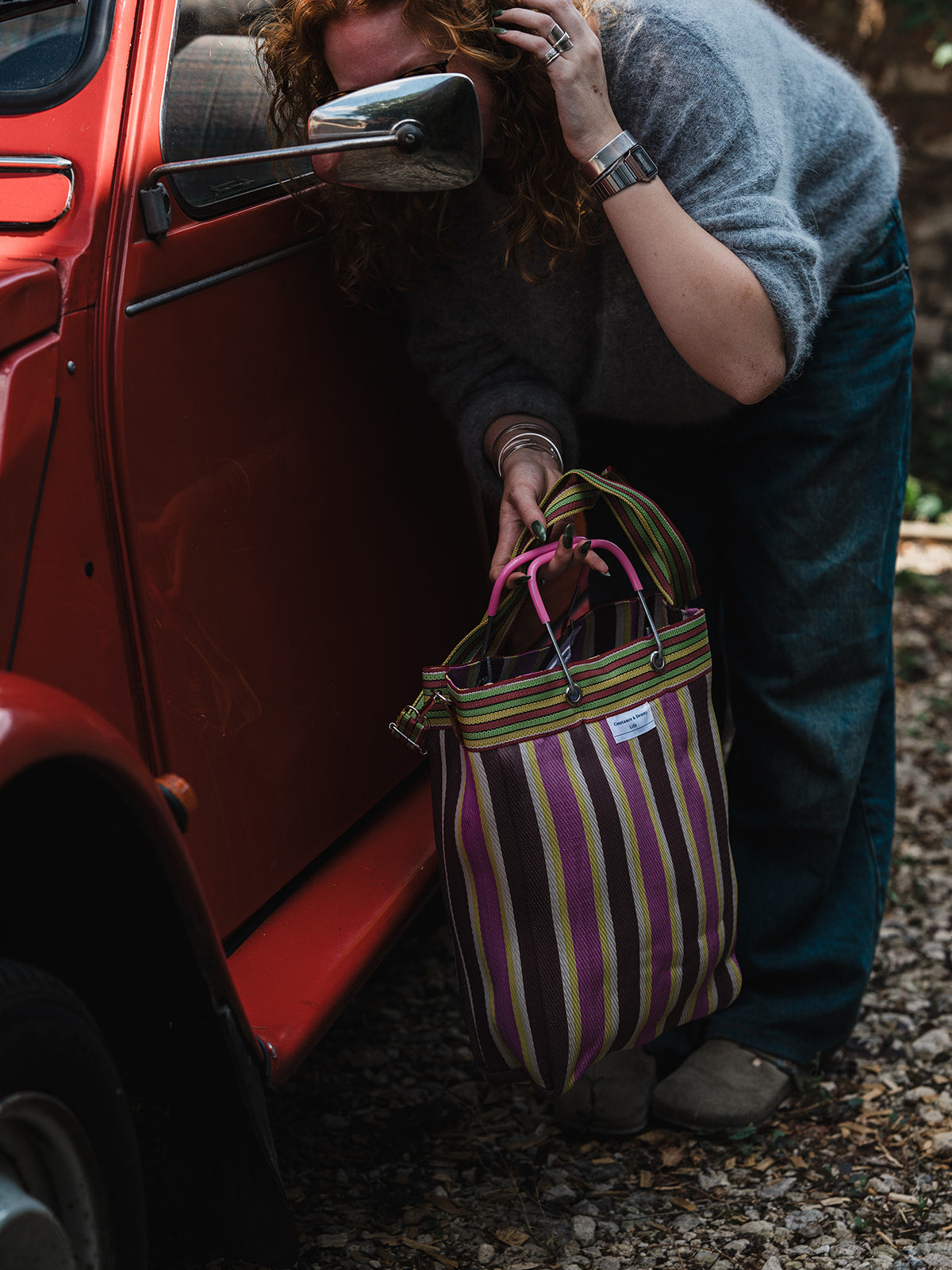 A person outdoors beside a red vintage car holds a striped tote bag with pink handles. The bag has vertical stripes in purple, green, beige, and brown, with a small white brand label near the top.