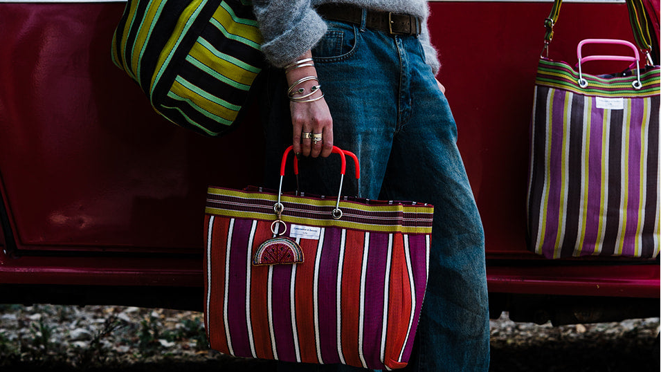 A person standing beside a red car holds a striped tote bag in front of them. Two matching striped tote bags are visible: one hanging on the car door and one being carried.