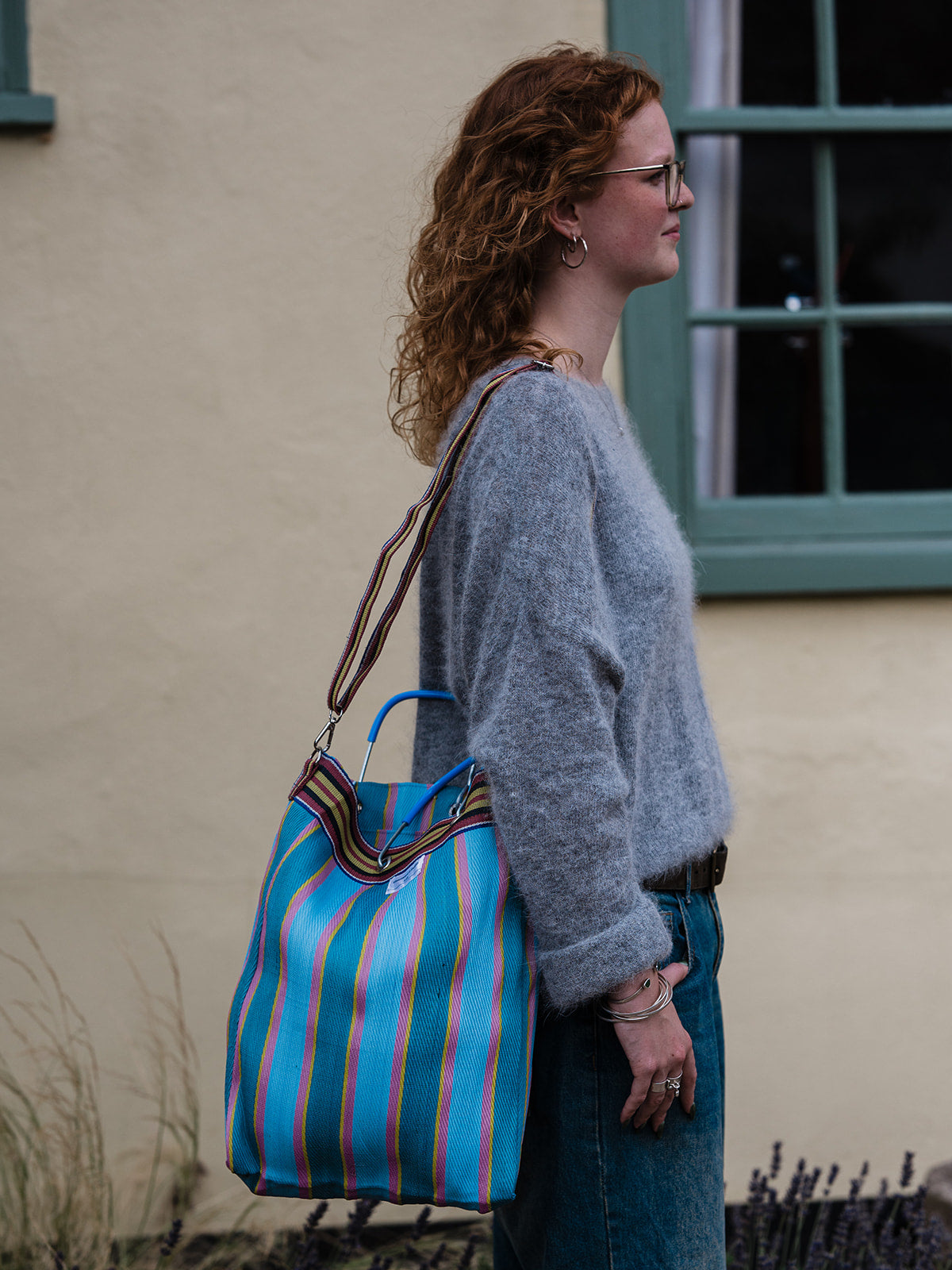 A woman with curly red hair wearing a gray knit sweater and blue jeans stands in profile outdoors, carrying a large striped tote bag with blue, teal, orange, and brown vertical stripes.