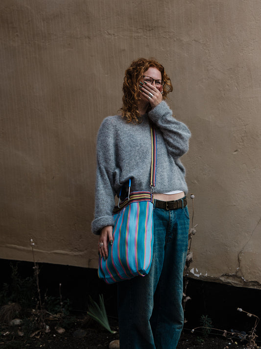 A person with curly red hair and glasses stands against a textured beige wall, wearing a gray knit sweater, blue jeans, and carrying a striped tote bag with blue, pink, and white vertical stripes.