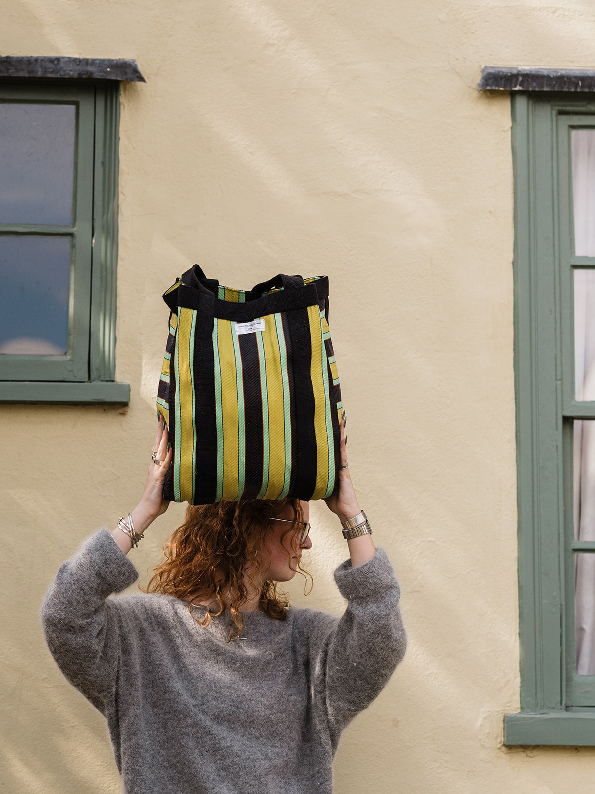 A person in a gray sweater balances a striped tote bag on their head against a beige stucco wall with green-trimmed windows.