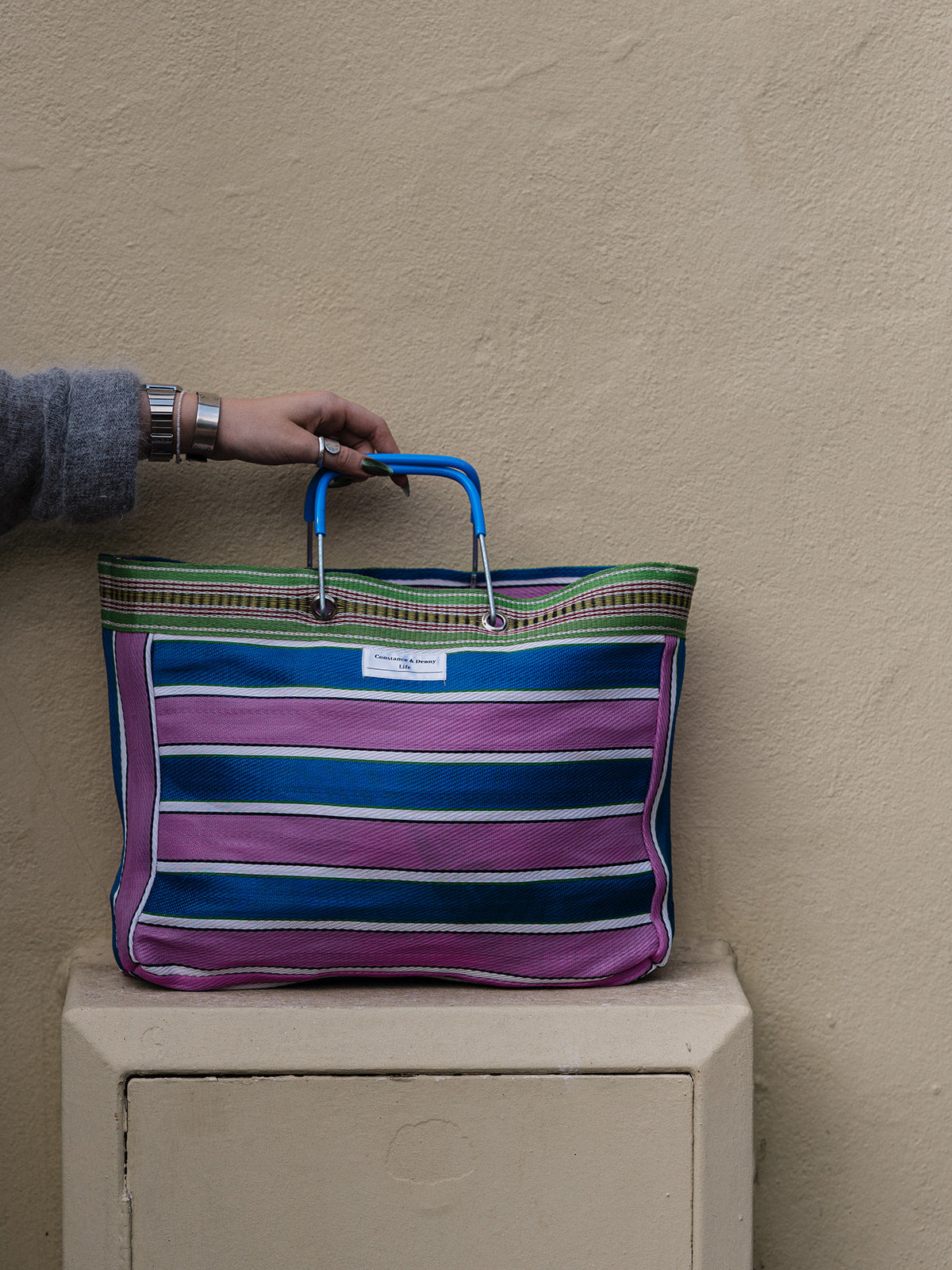 A striped tote bag with blue handles, featuring horizontal stripes in purple, blue, green, and white, resting on a beige stone pedestal against a textured beige wall. A hand wearing a gray sweater and silver bracelets holds the handle.