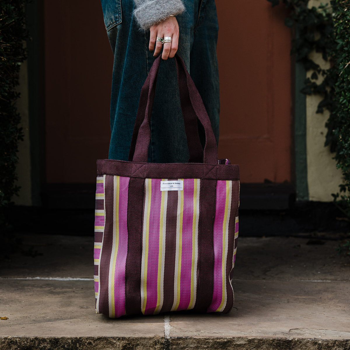 A striped canvas tote bag with pink, purple, brown, and cream vertical stripes, held by a person wearing a gray sweater and blue jeans in front of a rustic brown door.