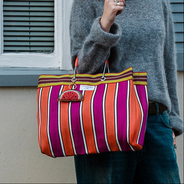 A woman with curly red hair and glasses stands outdoors against a cream wall with blue shutters, wearing a gray knit sweater, wide-leg blue jeans, and beige shoes while carrying a large striped tote bag with orange, pink, purple, and yellow vertical stripes.