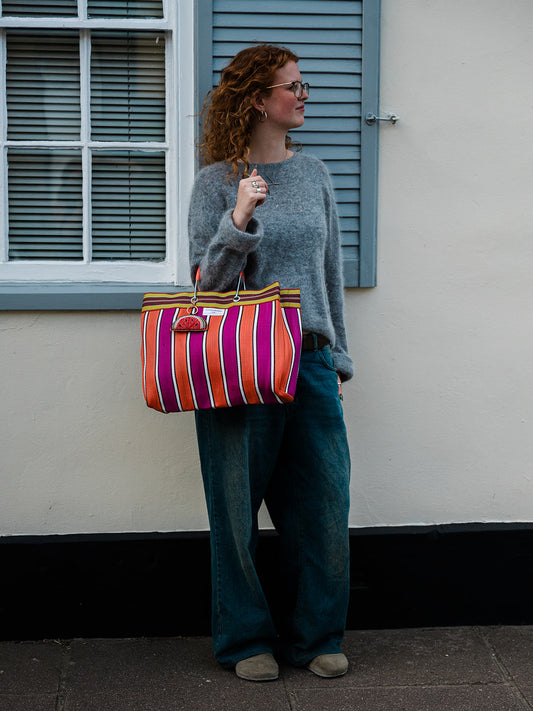 A woman with curly red hair and glasses stands outdoors against a cream wall with blue shutters, wearing a gray knit sweater, wide-leg blue jeans, and beige shoes while carrying a large striped tote bag with orange, pink, purple, and yellow vertical stripes.