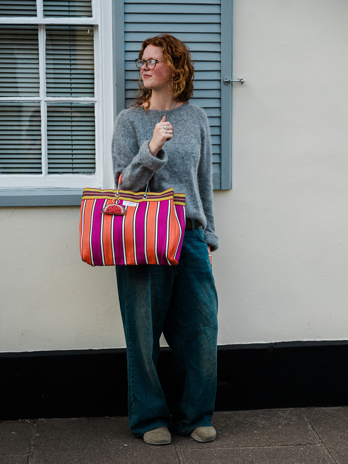 A woman with curly red hair and glasses stands outdoors against a cream-colored wall with blue shutters, wearing a gray knit sweater, wide-leg teal jeans, and beige shoes, while holding a colorful striped tote bag with pink, orange, and purple stripes.