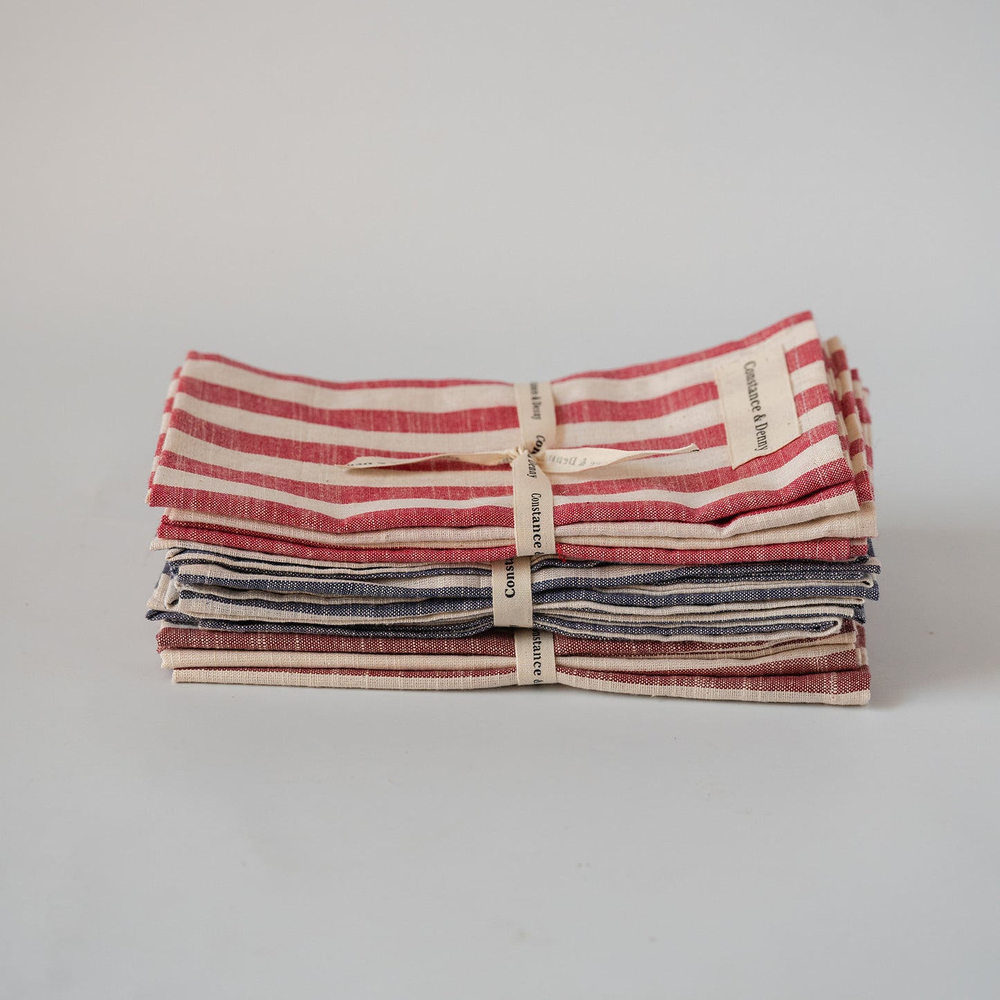 A stack of three folded striped linen napkins tied with a small ribbon. The top napkin has red and cream stripes, the middle has navy and cream stripes, and the bottom has dark red and cream stripes.