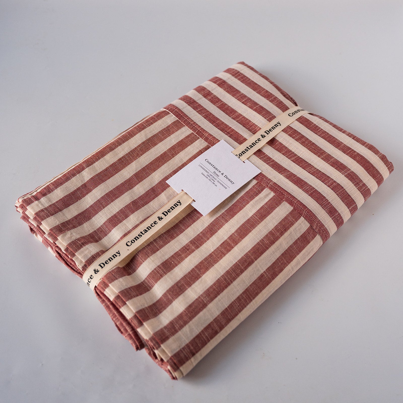 A folded striped textile, likely a blanket or throw, in muted terracotta red and cream stripes, wrapped with a paper band and tag. The brand 'Constance & Deny' is clearly printed on the band.