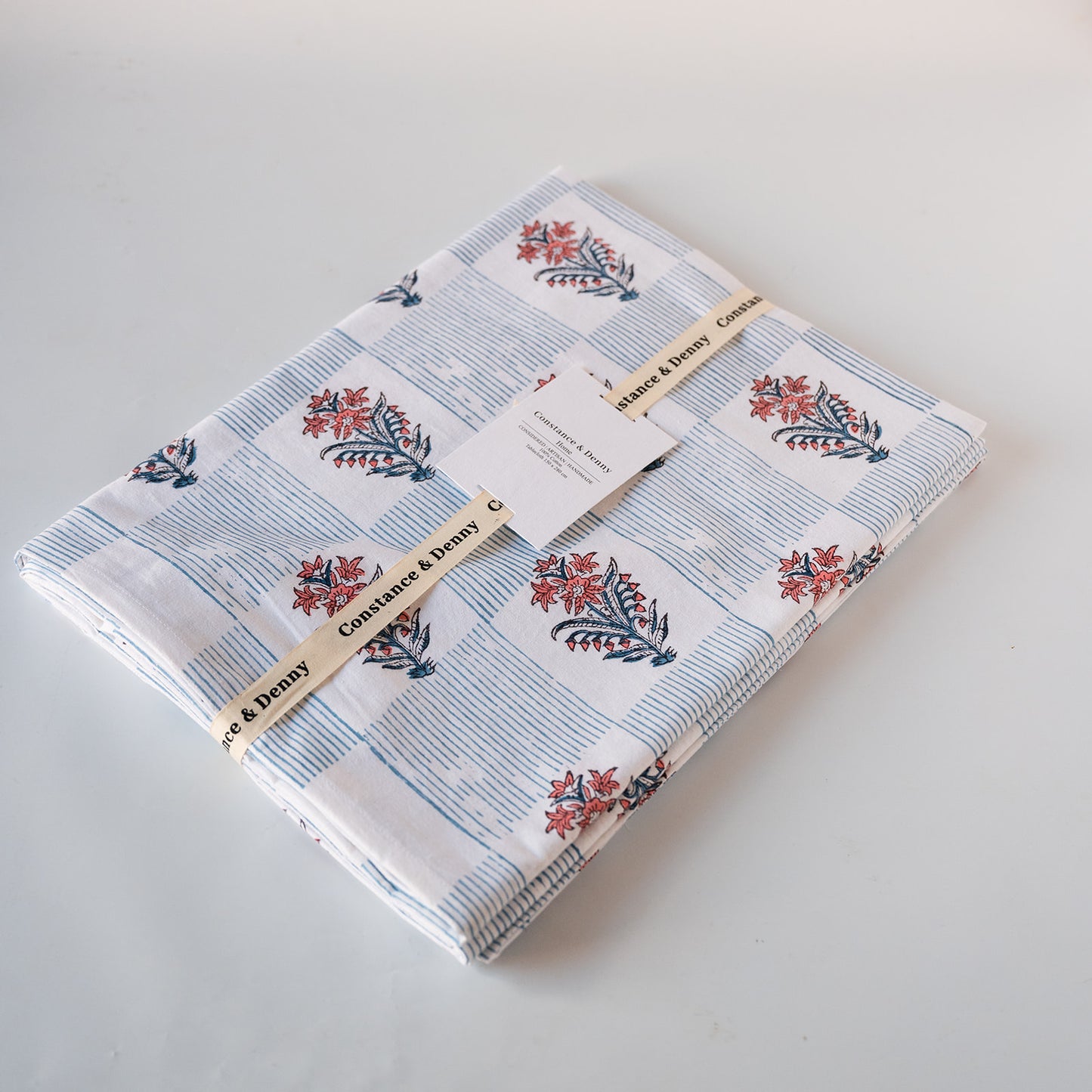 A folded cotton fabric bundle with a blue and white striped pattern and red floral motifs, wrapped with a paper band labeled 'Saffron & Danni' and a white product tag, on a light gray background.