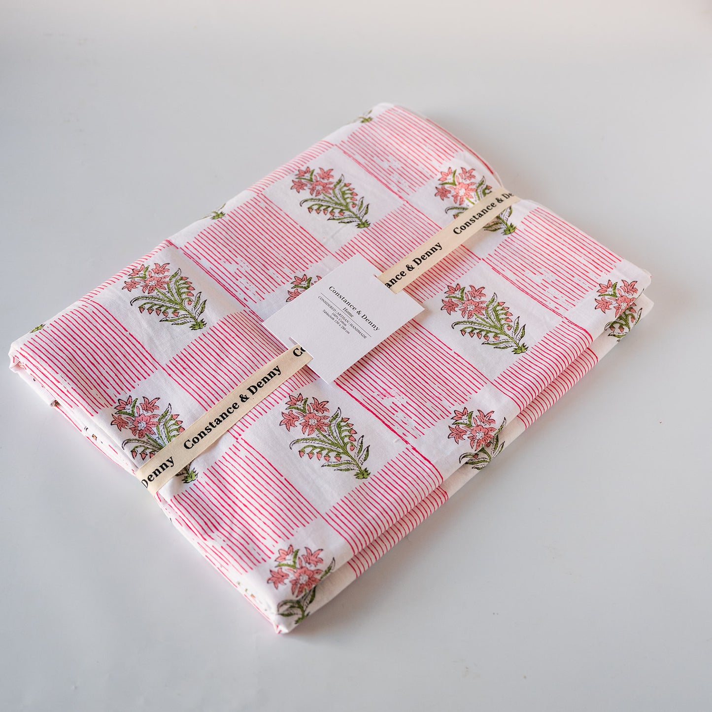 A folded cotton blanket with a pink and white checkered pattern, featuring pink floral prints with green leaves. The blanket is wrapped with a paper band labeled 'Rose & Daisy' and a white paper tag, photographed on a light gray background.
