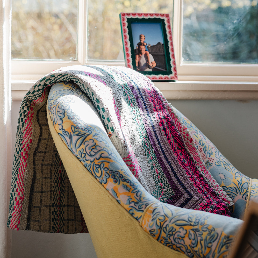 Colourful patterned blanket draped over a chair next to a window with a photo frame. Vintage-kantha-blanket-throw-textiles