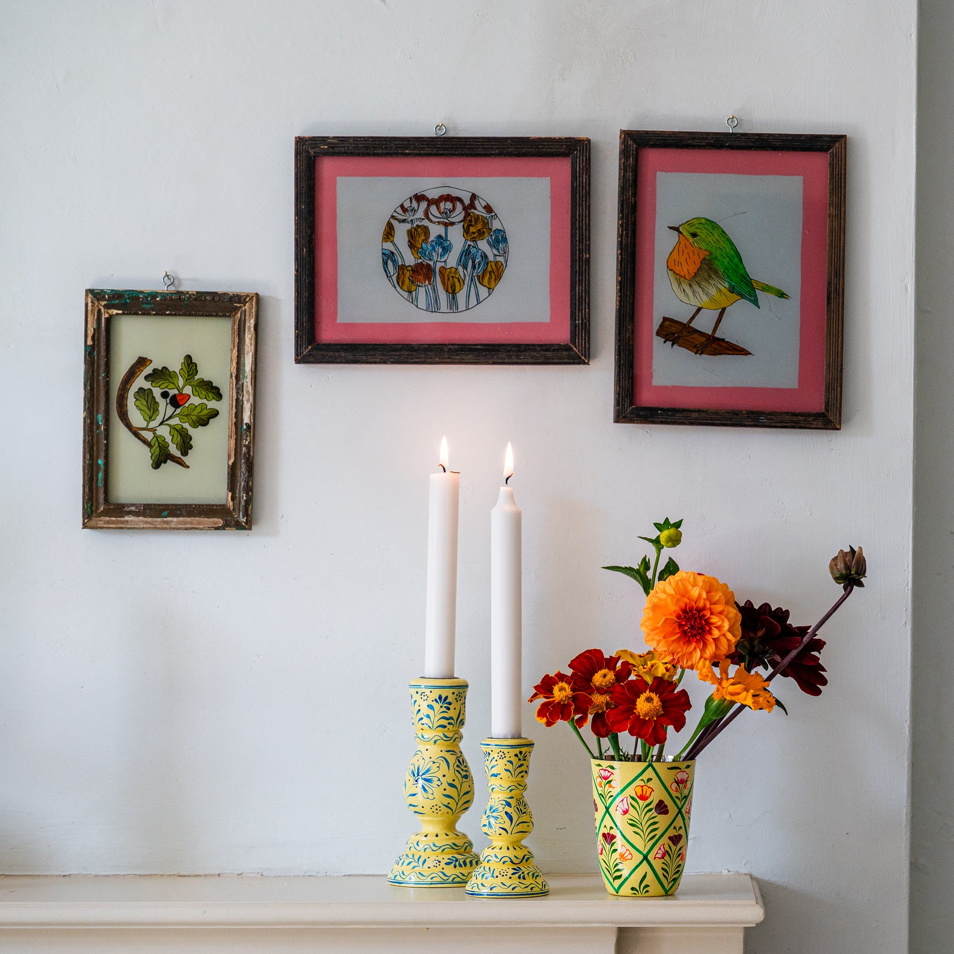 Decorative setup with framed artwork, candles, and a vase of flowers on a white surface.