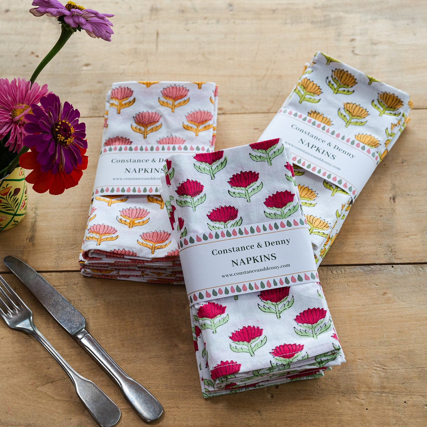 Set of floral-patterned napkins on a wooden table with cutlery and flowers.