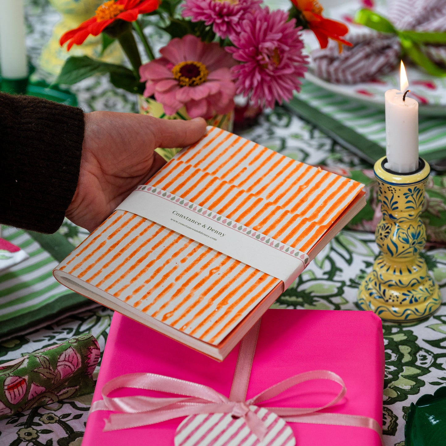 Person holding a striped notebook with a pink gift box and floral arrangement in the background