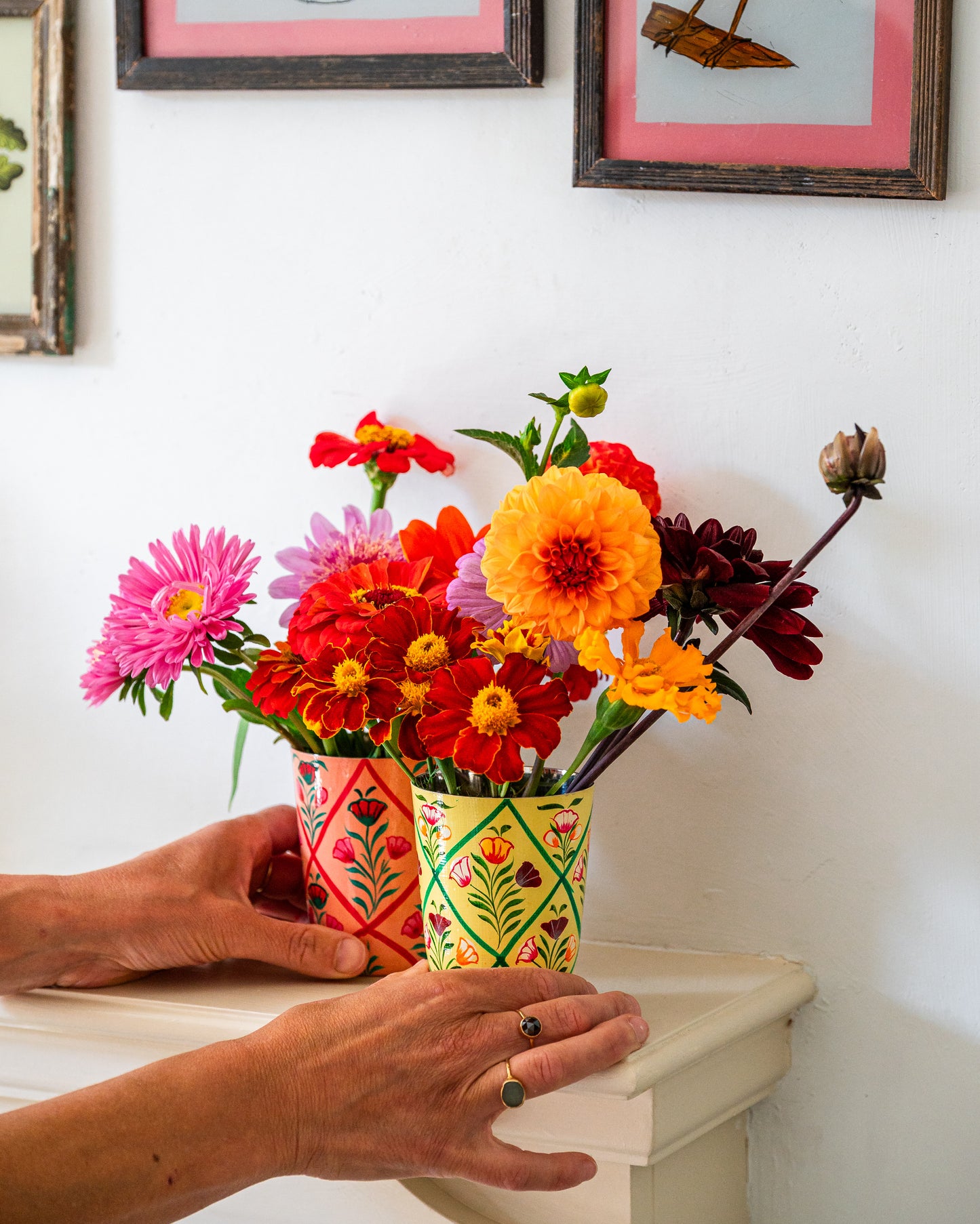 Colorful bouquet of flowers in a decorated vase held by hands against a white wall with framed pictures.