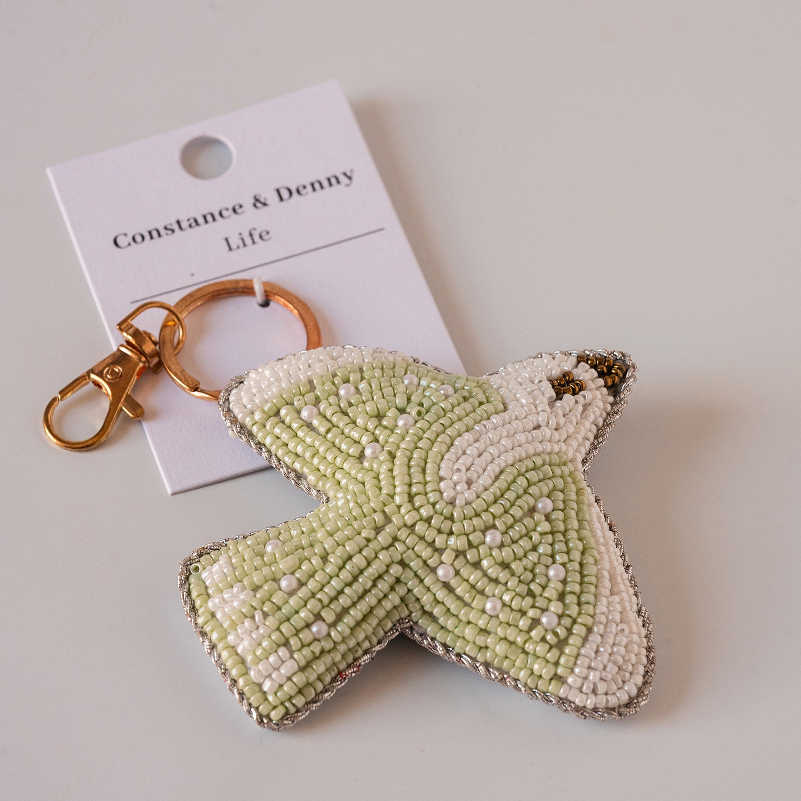 A handmade beaded bird keychain shaped like a dove, featuring light green and white seed beads with a gold-tone clasp, displayed next to its white paper tag that reads 'Constance & Denny Life'.
