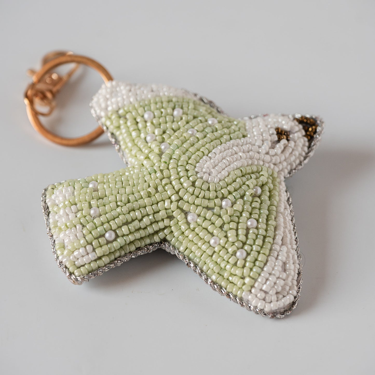 A handmade beaded bird keychain charm in pale green and white, featuring a gold-tone split ring, photographed on a neutral light gray background.