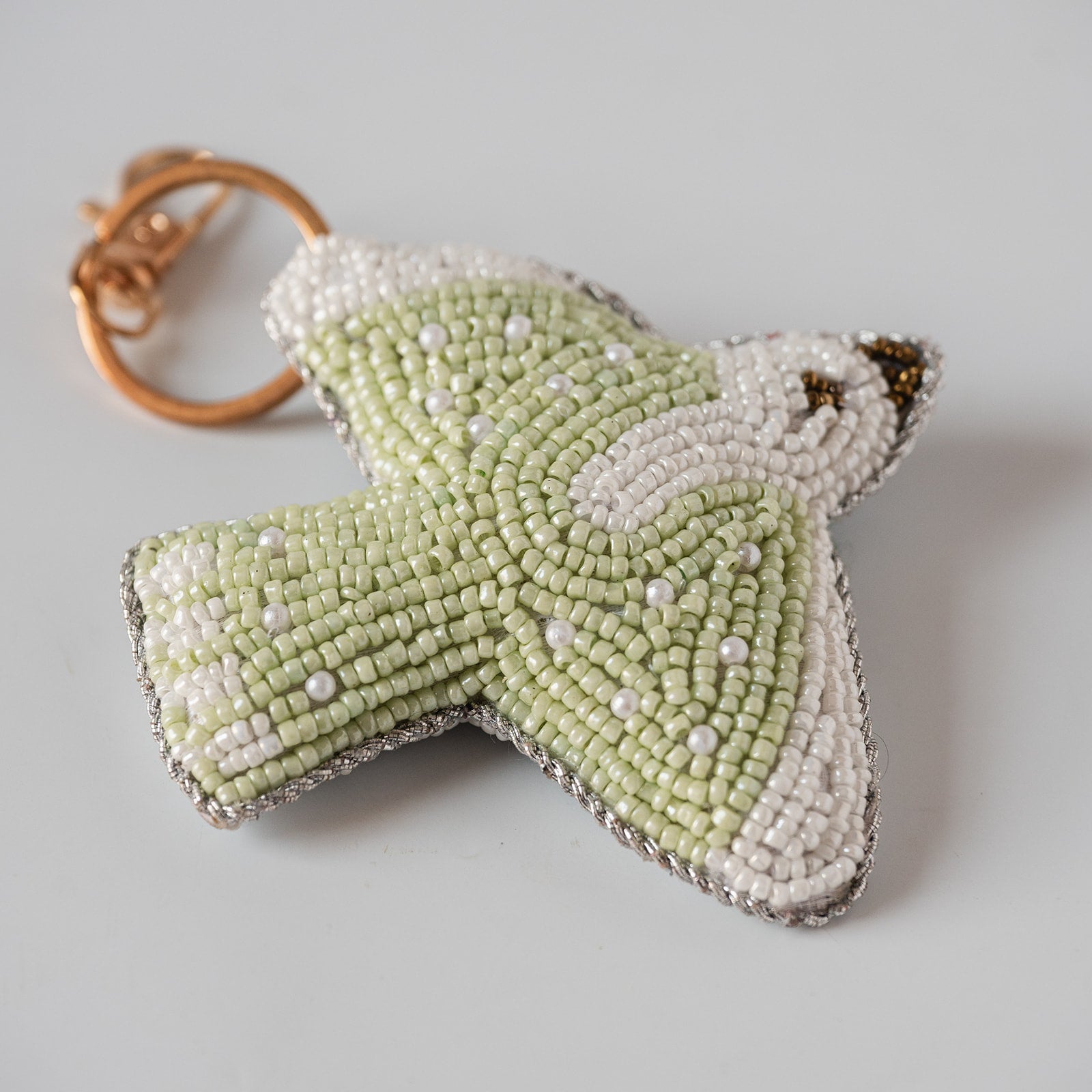 A handmade beaded bird keychain charm in pale green and white, featuring a gold-tone split ring, photographed on a neutral light gray background.