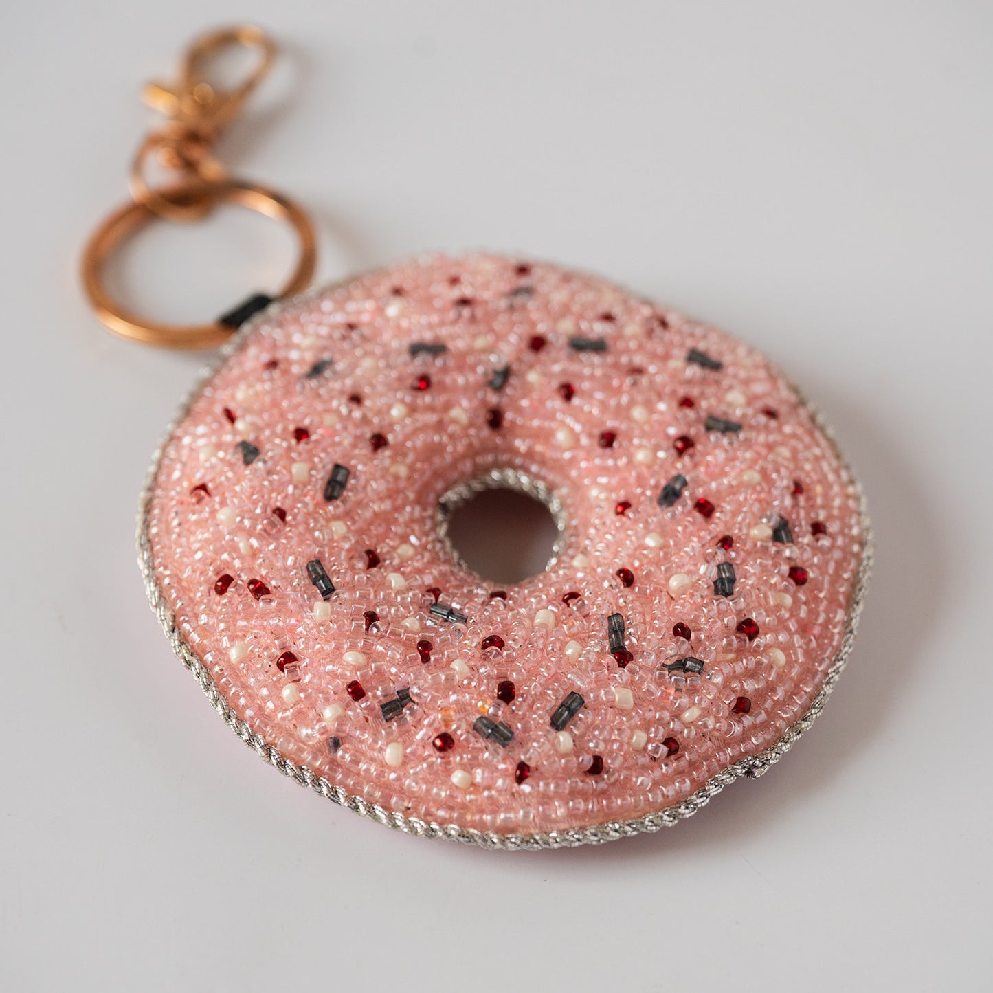 A pink donut-shaped keychain charm covered in white and red sprinkles, with a gold-tone split ring and clasp on a light gray background.