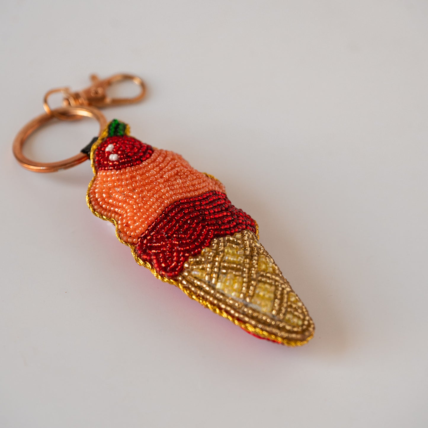 A beaded ice cream cone keychain with a copper-toned clasp, featuring orange and red beaded scoops, a yellow beaded waffle cone, and a small green top detail, shown on a plain light background.