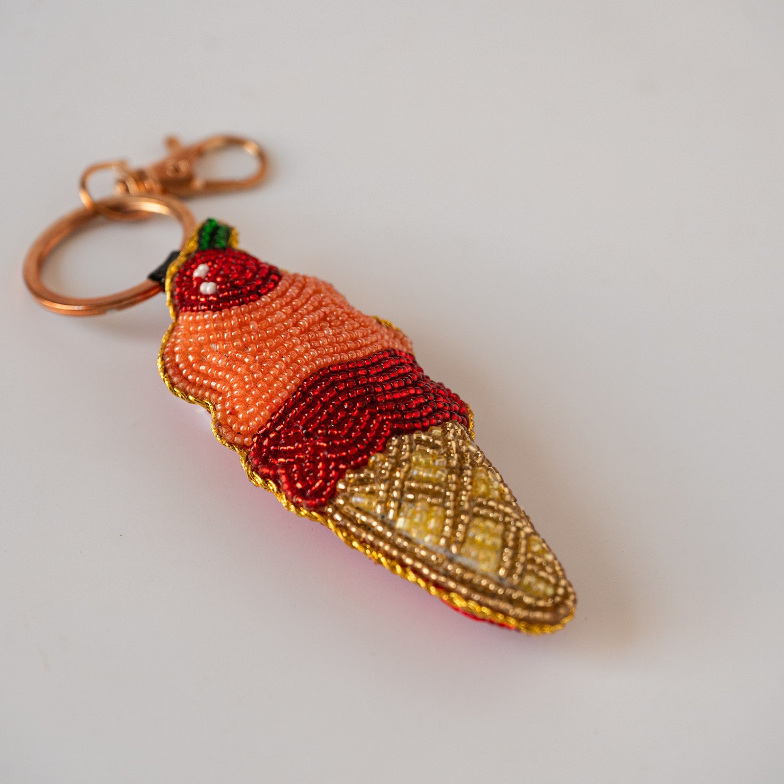 A beaded ice cream cone keychain with a copper-toned clasp, featuring orange and red beaded scoops, a yellow beaded waffle cone, and a small green top detail, shown on a plain light background.