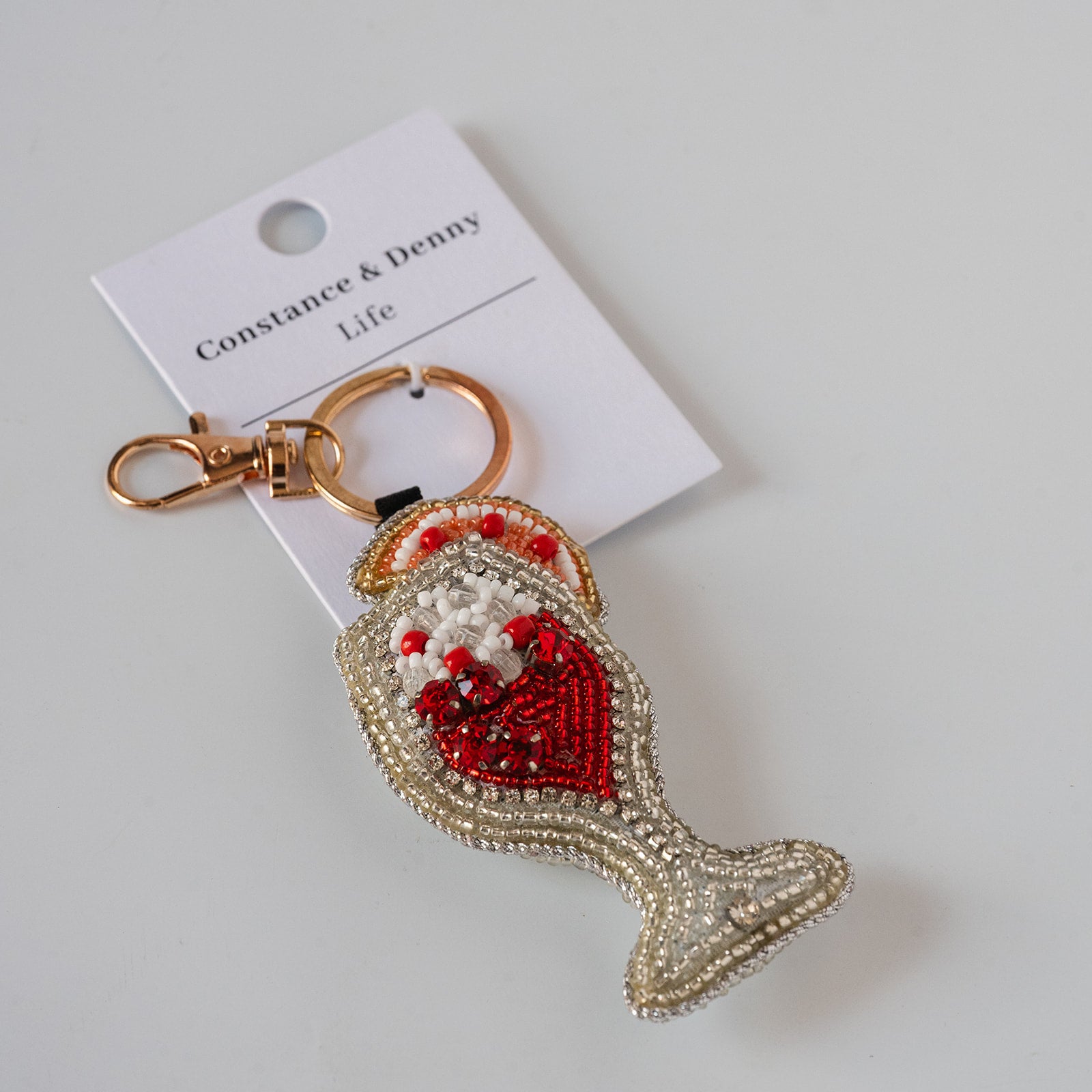 A fish-shaped keychain bag charm with a red and white beaded heart design, attached to a gold-tone clasp and displayed on a white card labeled 'Constance & Denny Life' against a light gray background.