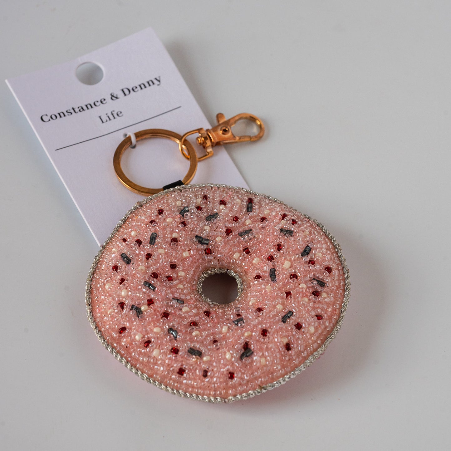 A pink beaded donut keychain with gold-tone hardware and a white paper tag reading 'Constance & Denny Life', photographed on a light neutral background.
