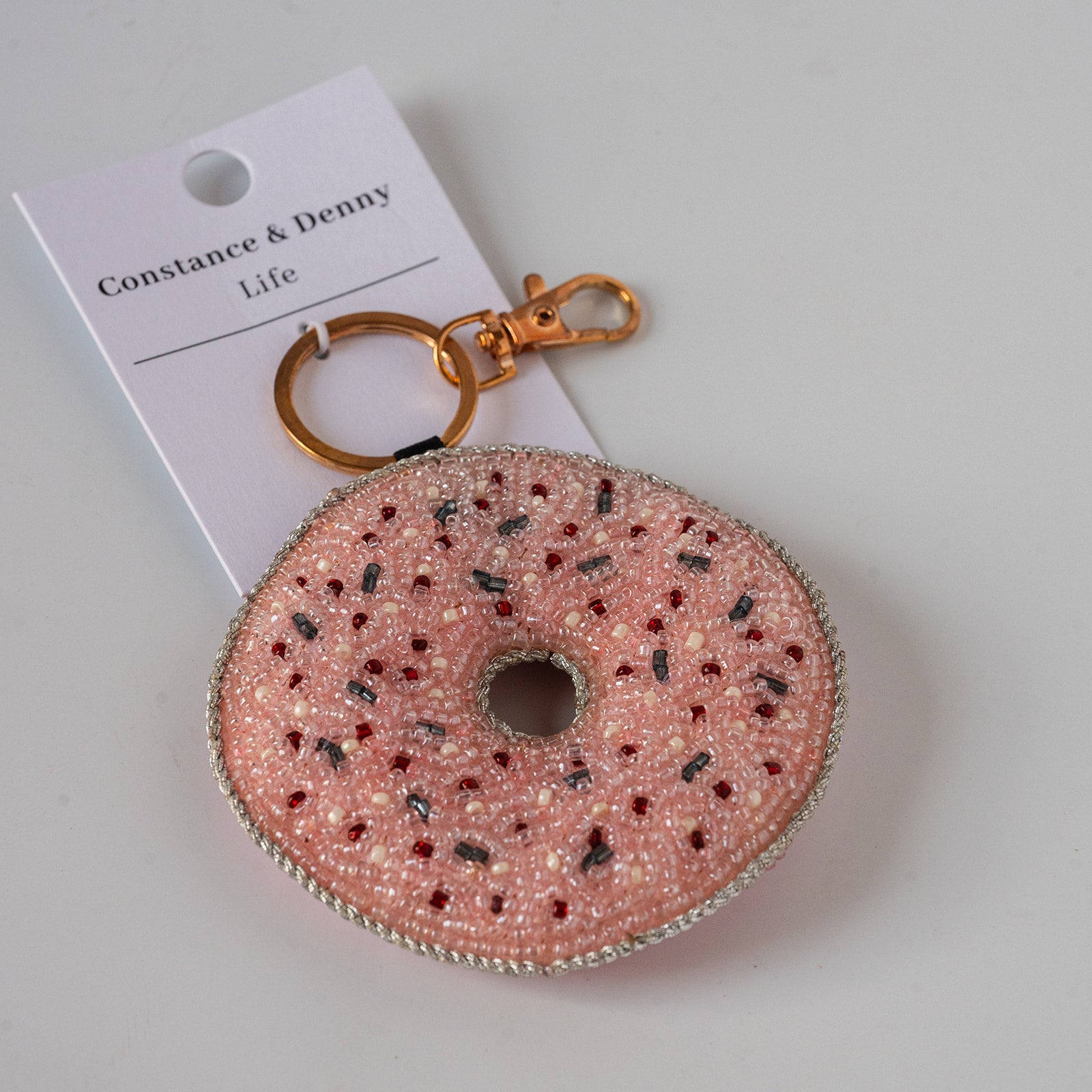 A pink beaded donut keychain with gold-tone hardware and a white paper tag reading 'Constance & Denny Life', photographed on a light neutral background.