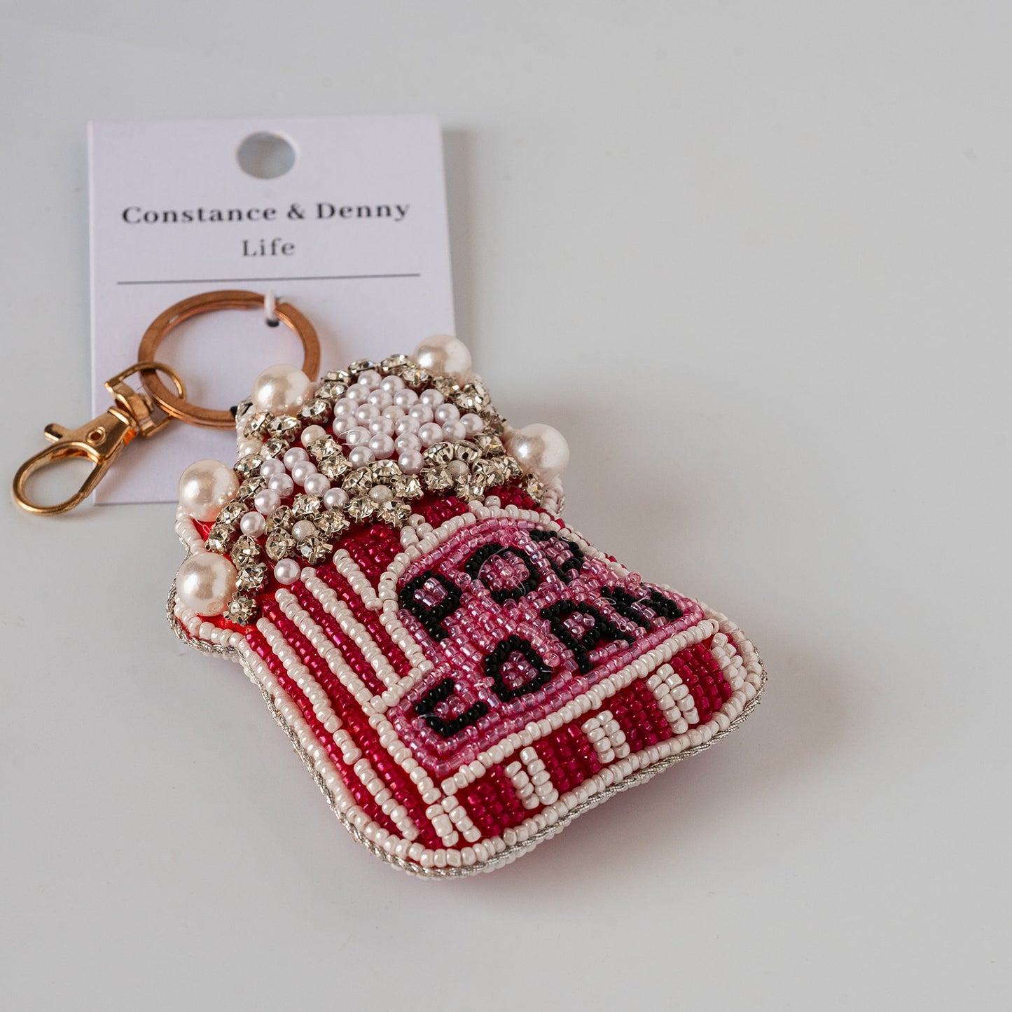 A beaded keychain pouch shaped like a red and pink striped popcorn bag, decorated with faux pearls and rhinestones, attached to a gold-tone clasp and a white Constance & Denny Life brand card.