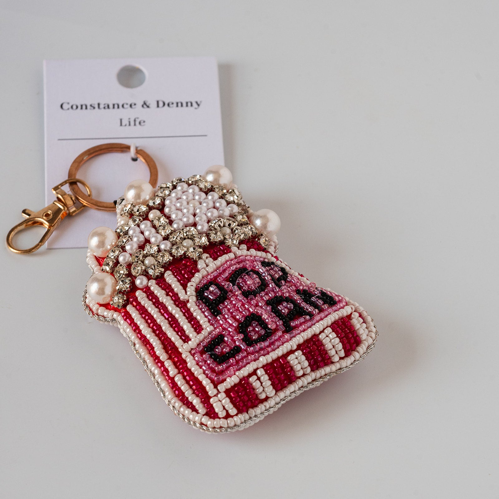 A beaded keychain pouch shaped like a red and pink striped popcorn bag, decorated with faux pearls and rhinestones, attached to a gold-tone clasp and a white Constance & Denny Life brand card.