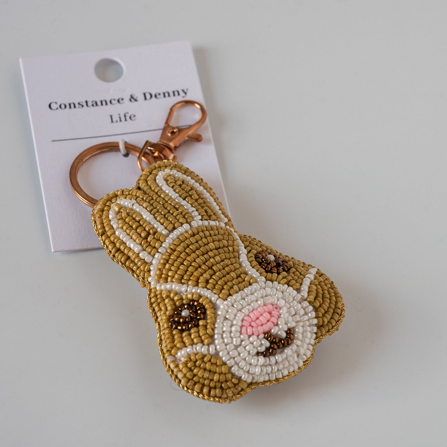 A beaded bunny-shaped keychain attached to a white card labeled 'Constance & Denny Life', photographed on a light gray background.