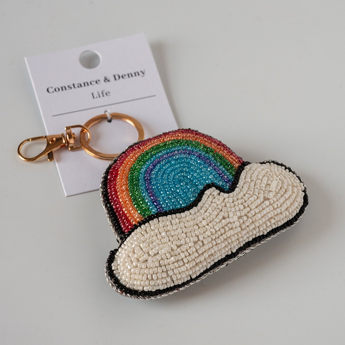 A beaded rainbow cloud keychain with a gold clasp, shown with its white paper hangtag that reads 'Constance & Denny Life' on a light gray background.
