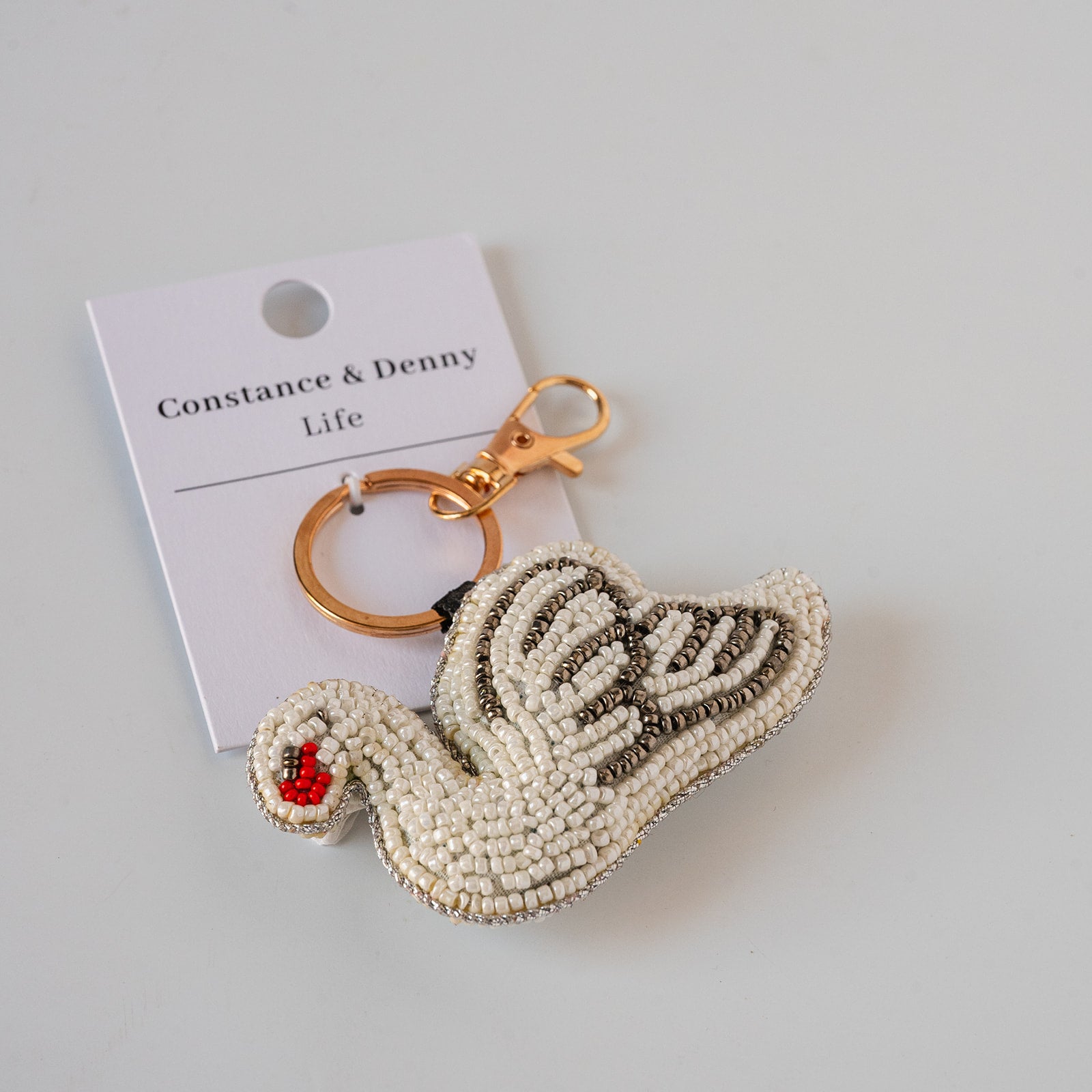 A swan-shaped beaded keychain with a gold clasp, displayed on its white brand card which reads 'Constance & Denny Life'. The swan is primarily cream and white beaded with black detailing, a red beaded beak, and a small red ladybug accent.