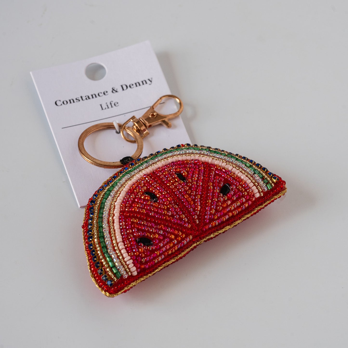 A watermelon slice keychain charm made of red, green, and cream beaded segments, attached to a white paper tag that reads 'Constance & Denny Life' and featuring a gold-tone lobster clasp, on a light gray background.