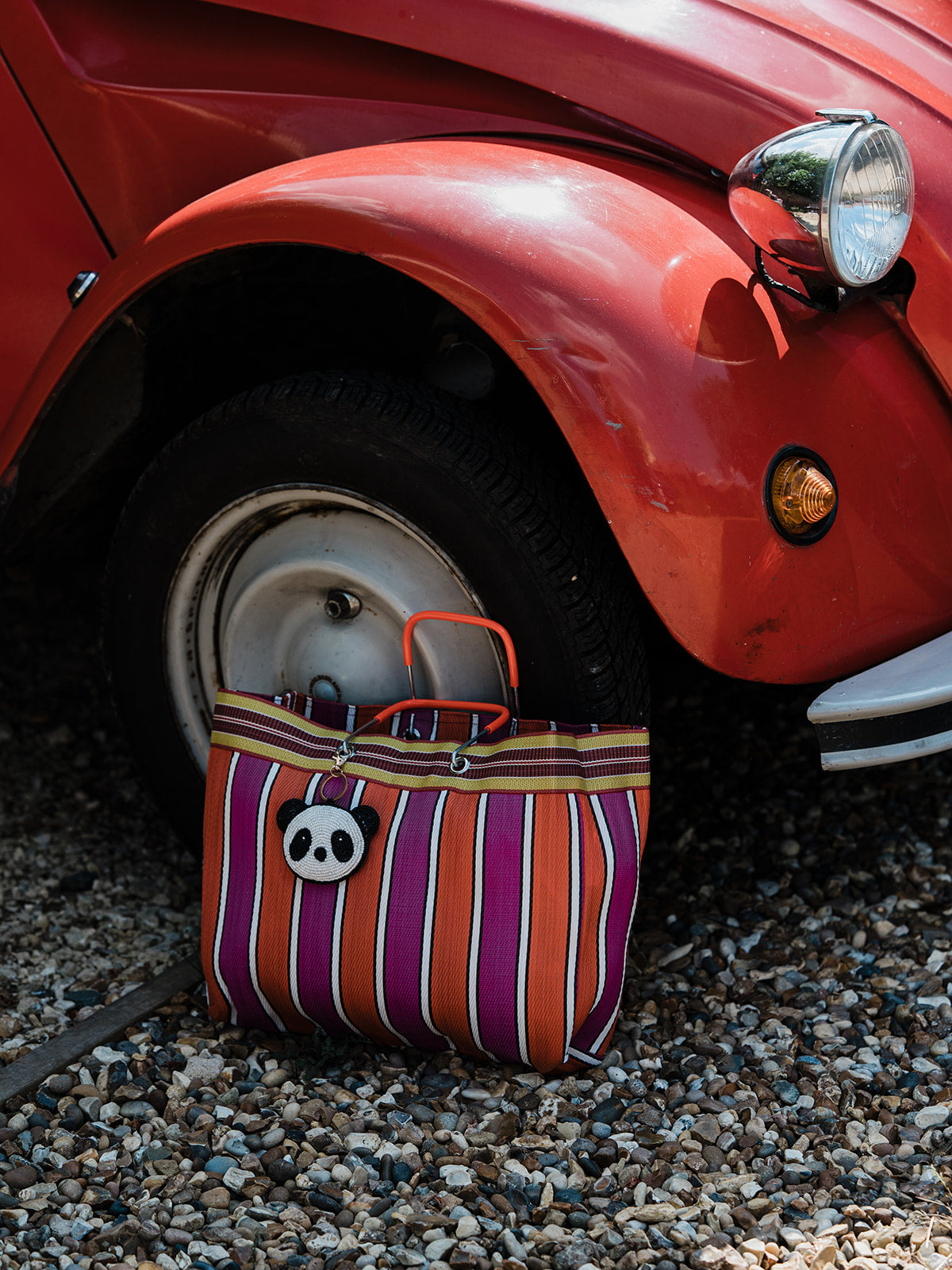 A striped tote bag with purple, orange, and white vertical stripes, orange handles, and a white panda face charm, resting on gravel next to the front wheel of a red car.