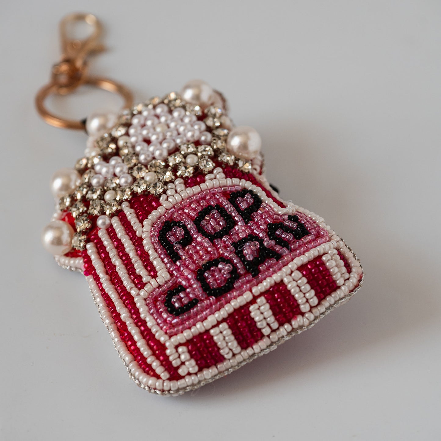 A beaded popcorn-shaped keychain charm with a gold clasp, featuring red and white striped sides, a pink top, white pearl-like beads, and black embroidered text reading 'POP CORN' on a light gray background.
