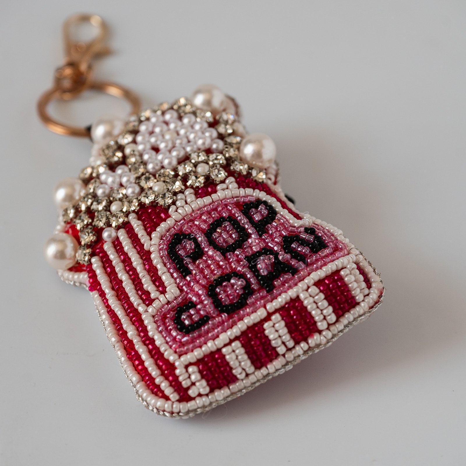 A beaded popcorn-shaped keychain charm with a gold clasp, featuring red and white striped sides, a pink top, white pearl-like beads, and black embroidered text reading 'POP CORN' on a light gray background.