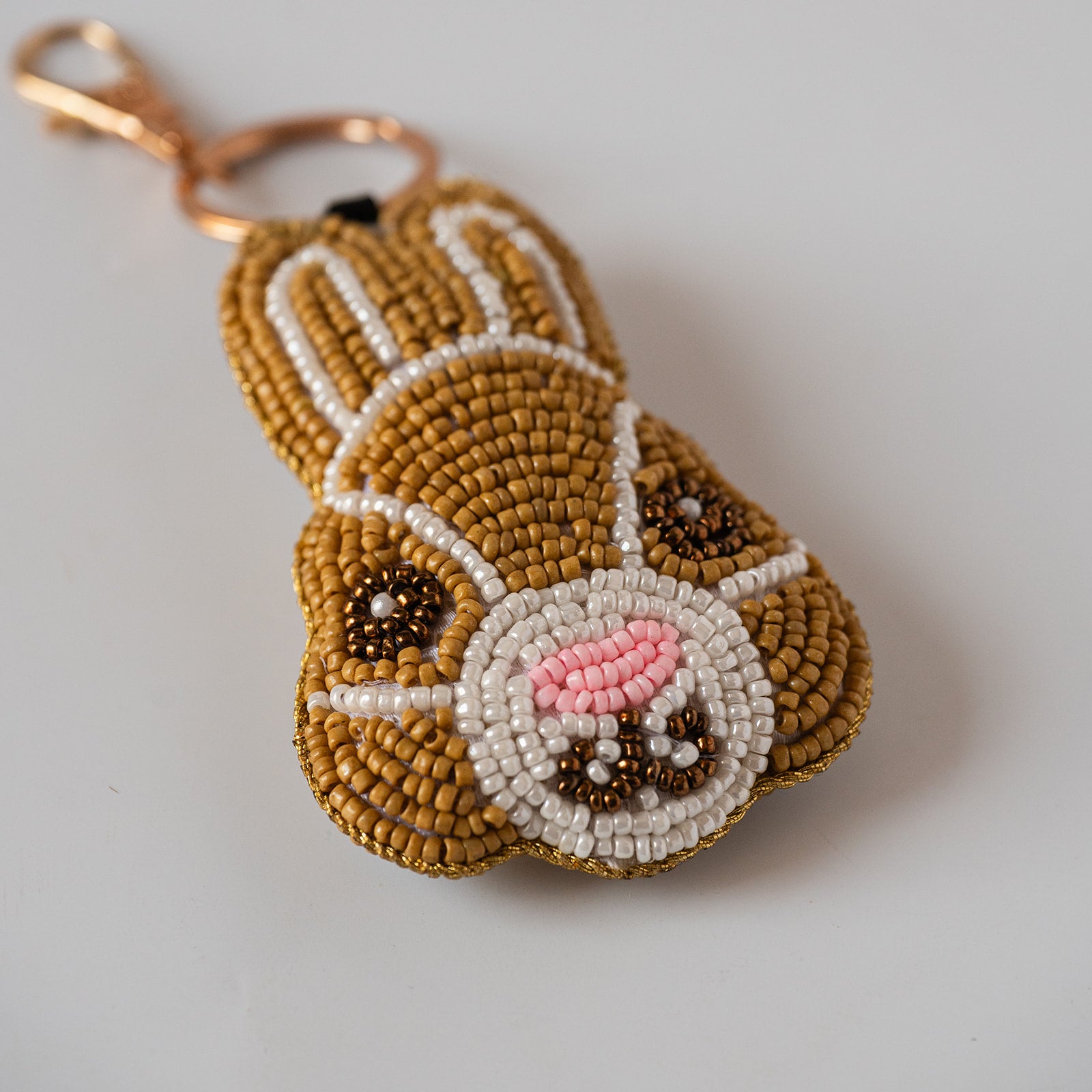 A handmade beaded keychain shaped like a bunny face, featuring tan, white, pink, and brown seed beads with a gold-tone lobster clasp, shown on a light gray background.