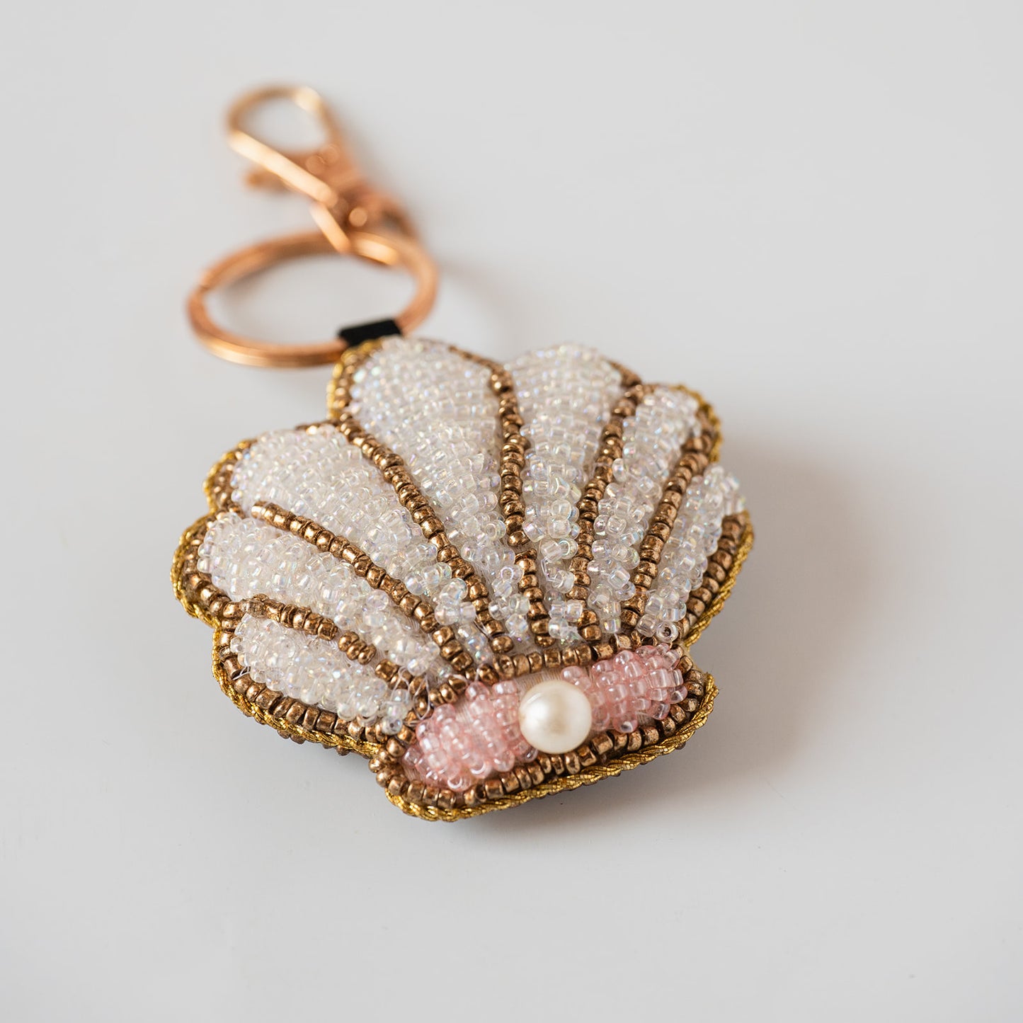 A handmade seashell-shaped keychain or bag charm made of white and pink seed beads with gold metallic trim and a central pearl, attached to a rose gold clasp on a light gray background.