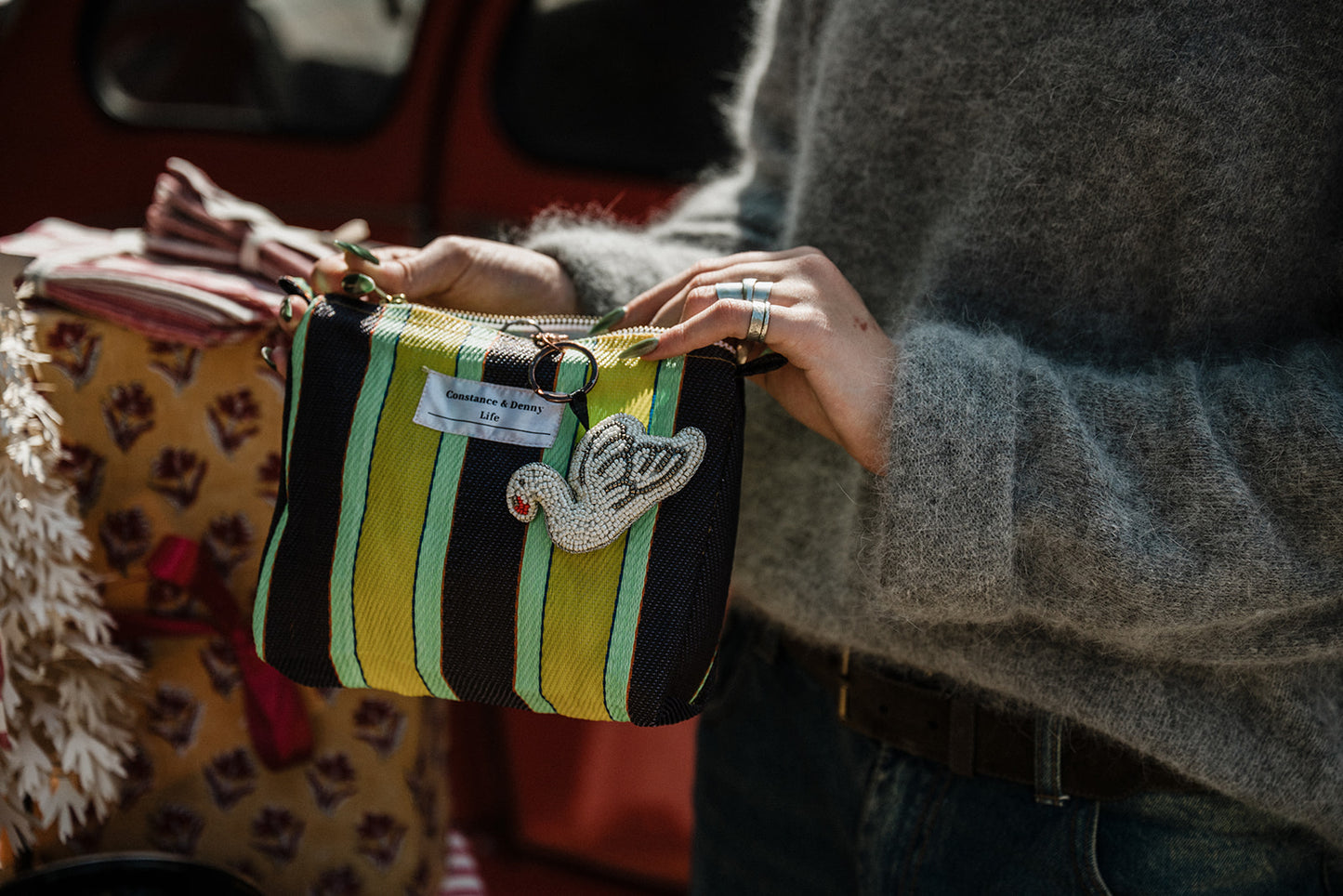 A close-up of a person carrying a striped pouch bag outdoors. The bag has vertical stripes in black, green and yellow, with a decorative embroidered swan shaped charm.