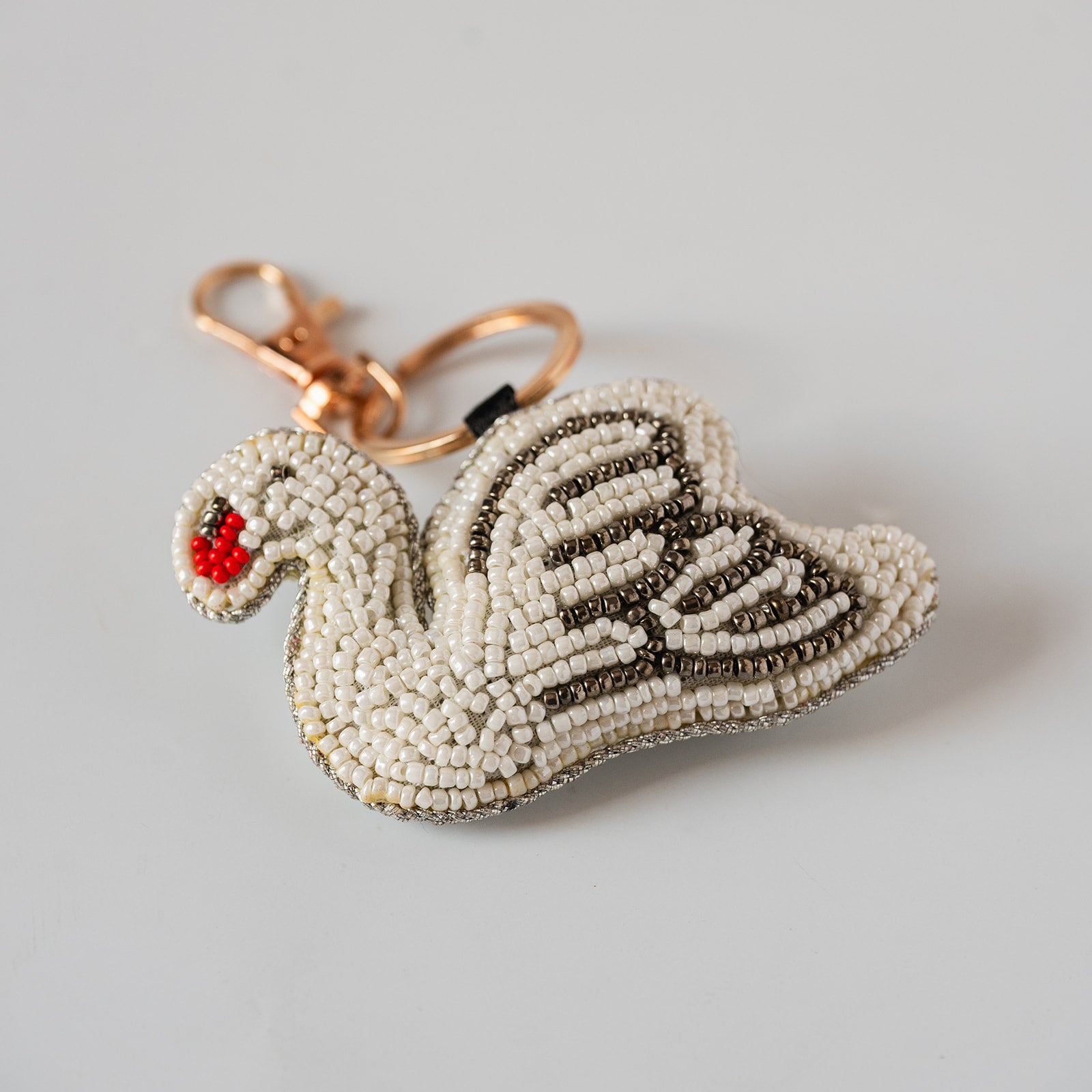 A swan-shaped beaded keychain with a white and brown bead body, a small red and green beaded beak, and a rose gold metal clasp on a light gray background.