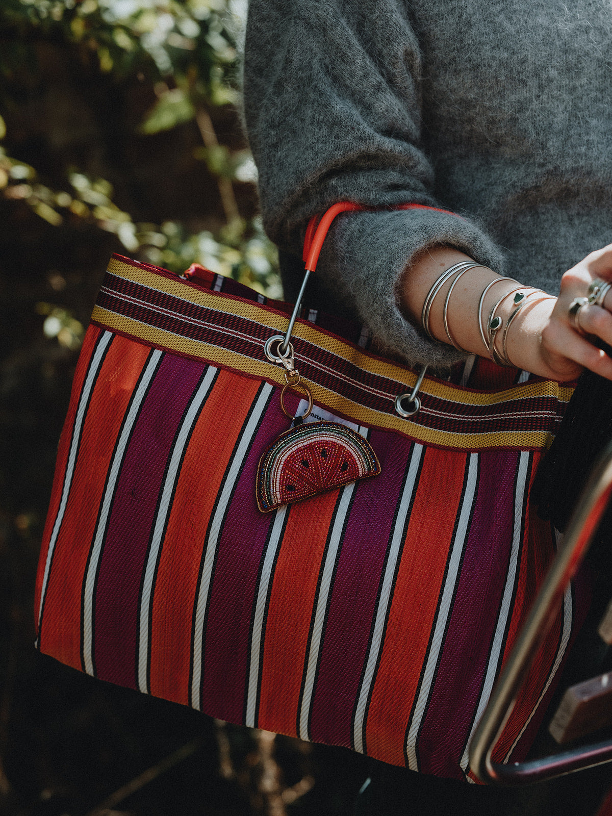 A close-up of a person carrying a large striped tote bag outdoors. The bag has vertical stripes in red, orange, purple, and white, with a decorative embroidered watermelon shaped charm.